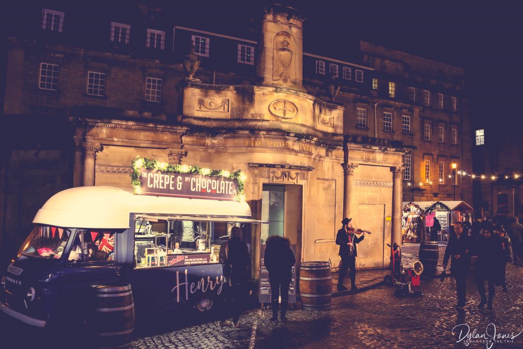 A Crepe and Chocolate Van and a violinist outside of The Cross Bath at the Bath Christmas Market